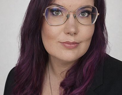 Professionelles Bewerbungsfoto einer Frau mit schulterlangem, violettem Haar und Brille, frontal in die Kamera blickend. Ruhiger, neutraler Gesichtsausdruck, dezentes Make-up, Septum-Piercing. Oberkörperporträt vor hellem, neutralem Studiohintergrund mit gleichmäßiger Ausleuchtung.