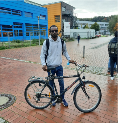 Ein Student mit Fahrrad auf einem gepflasterten Campusplatz vor modernen Universitätsgebäuden.