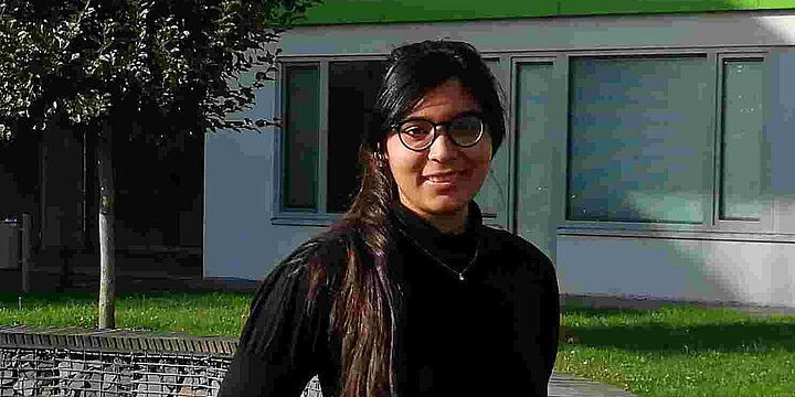 Automatisierungstechnik Junge Frau mit dunklen Haaren und schwarzem Pulli steht auf einer grünen Wiese vor einem Gebäude