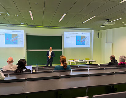 In diesem Foto sieht man wie Menschen einem Vortrag folgen der von einer Person gehalten wird.