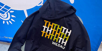 dunkelblauer Hoodie mit weiß-grün-orangefarbenem Muster vor einer blau-weißen Logowand