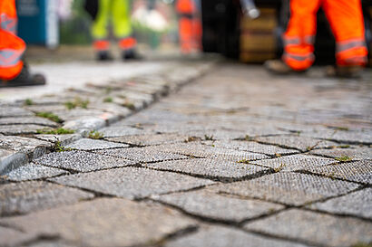 Detailaufnahme eines Kopfsteinpflasters während Bauarbeiten zur Verbesserung der Fahrqualität für den Radverkehr, im Hintergrund sind unscharf Arbeiter in Warnschutzkleidung zu erkennen.