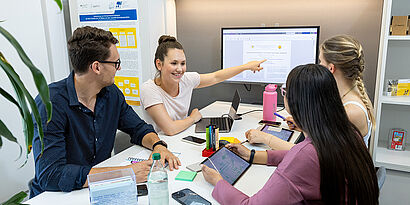 Studierende sitzen an einem Tisch und sind im Gespräch, eine Studierende zeigt auf einen Bildschirm.