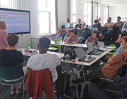 Studierende sitzen um einen Tisch, ein Professor hält einen Vortrag