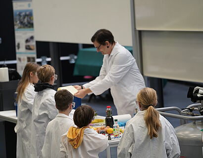 Kinderuni Winter Edition 2024 - Prof. Dreyer erklärt mehreren Kindern in Schutzkleidung ein anstehendes Experiment