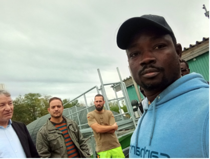 Eine Gruppe von vier Männern macht ein Selfie auf einem Gelände mit technischen Bauten im Hintergrund.