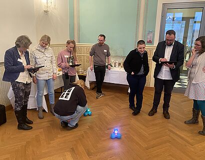 Hier sind ein paar Menschen zu sehen, die in einer Art Halbkreis stehen und kleine blaue Roboter auf dem Boden betrachten.