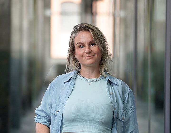 Portraitbild einer blonden Frau mit hellblauem Oberteil und blauem Hemd in einer Halle der TH Wildau