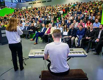Zwei Musiker*innen von hinten fotografiert, voll besetztes Audimax im Hintergrund