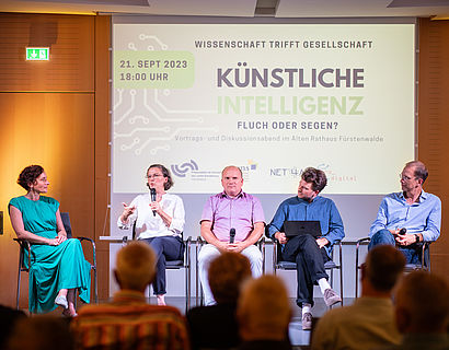 Personen sitzen in einer Gesprächsrunde zusammen und im Hintergrund hängt ein Screen mit der Aufschrift "Künstliche Intelligenz"