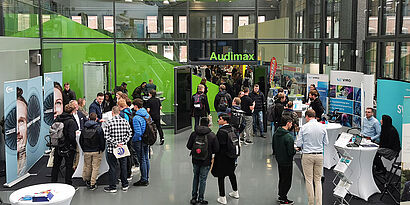 Besucherinnen und Besucher bei einer Messe an verschiedenen Unternehmenstischen im Gespräch