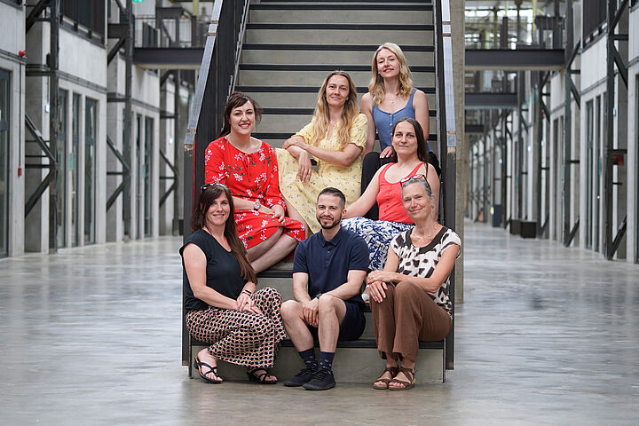 Sieben Personen sitzen für ein Gruppenbild auf einer Treppe und schauen in die Kamera