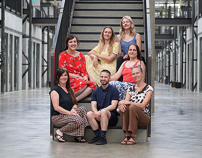 Sieben Personen sitzen für ein Gruppenbild auf einer Treppe und schauen in die Kamera