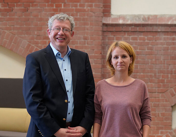 Zwei Personen, Mann links und Frau rechts, stehen vor einer roten Backsteinwand