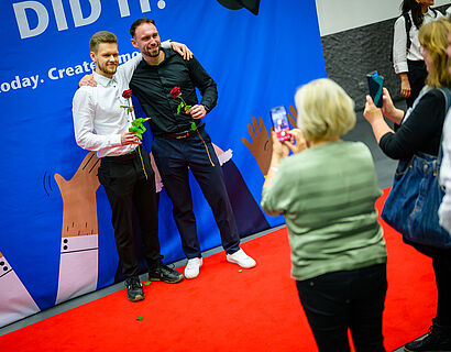 Zwei Absolventen stehen Arm in Arm für ein Foto beisammen und lächeln in die Kamera