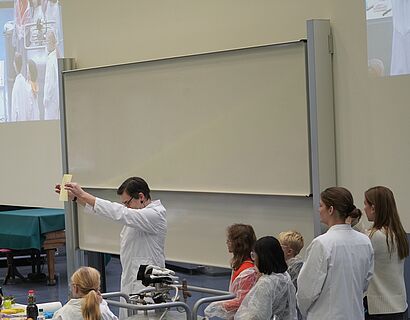 Kinderuni Winter Edition 2024 - Prof. Dreyer experimentiert, Kinder in weißer Schutzkleidung schauen gespannt zu 