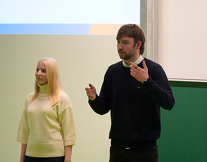 Teilnehmer*innen des THtalk, zwei Personen in eine Hörsaal vorne an der Tafel, die eine Person spricht und gestikuliert.