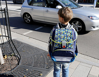 Kind mit Schulrucksack auf dem Rücken steht an einer Bordsteinkante und ein Auto fährt vorbei