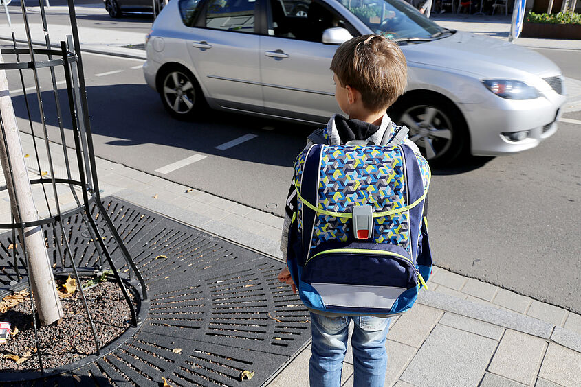Kind mit Schulrucksack auf dem Rücken steht an einer Bordsteinkante und ein Auto fährt vorbei