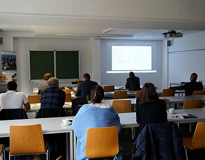 Hier ist ein Lehrraum zu sehen, mit Menschen die an Tischen sitzen und auf die Wand schauen wo eine PowerPoint dran projiziert wurde.