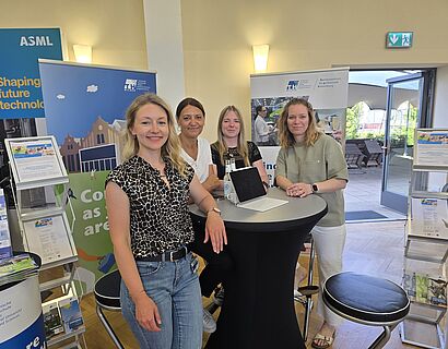 Vier Mitarbeiterinnen des KpE am stehen am Messestand und lachen in die Kamera, im Hintergrund sind Messematerialien zu sehen.