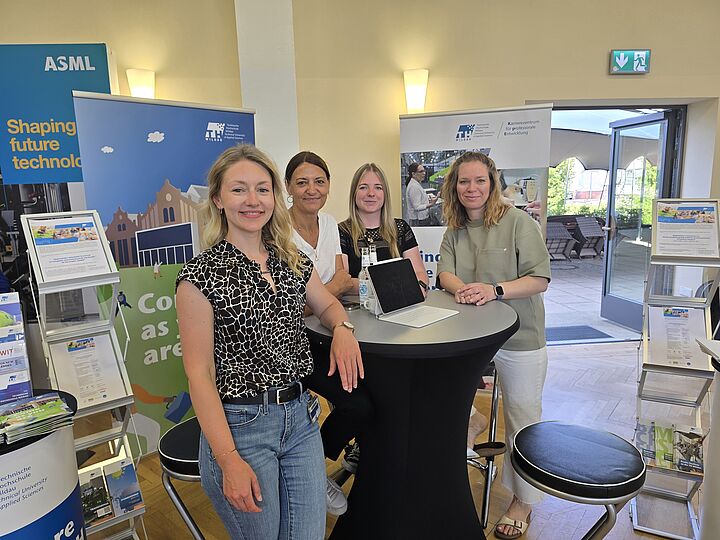 Vier Mitarbeiterinnen des KpE am stehen am Messestand und lachen in die Kamera, im Hintergrund sind Messematerialien zu sehen.