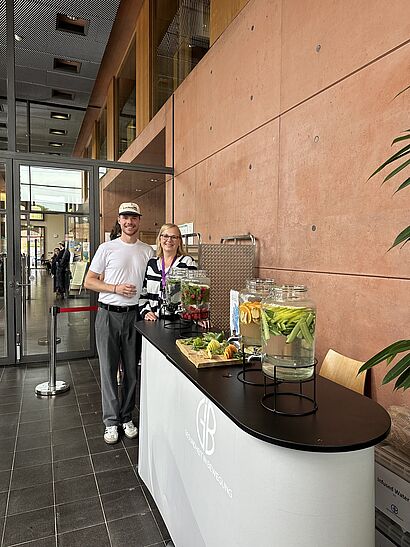 Max Rau (Gesundheitsbotschafter) und Monique Zamaitat (Hochschulberaterin der TK) stehen hinter einem weißen, rundlichen Stand mit großen Glasgefäßen voll Infused Water. Im Hintergrund ist die Fassade von Halle 14 mit Glastüren zu sehen.