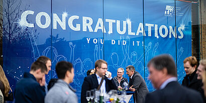 Blaues Banner mit Schriftzug "Congratulations" und Absolvent*innen im Vordergrund