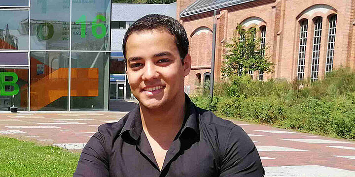 Automatisierungstechnik Junger Mann sitzt auf einer Bank auf dem Campus mit roten Backsteingebäuden im Hintergrund