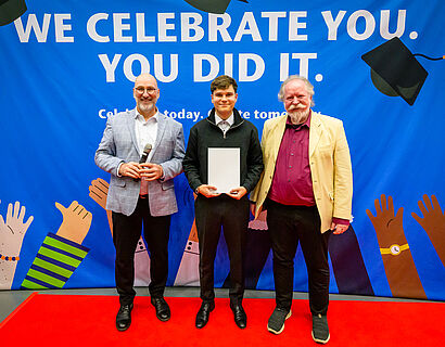 Drei Personen stehen vor einer Plakatwand, die mittlere Person hat eine Urkunde in den Händen