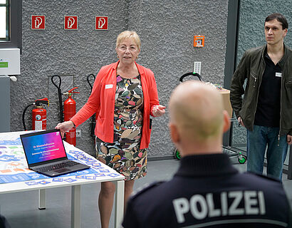 In diesem Bild ist eine Frau zu sehen, die ihre eine Hand am Computer hat, worauf eine Präsentation für die Veranstaltung zu sehen ist. Dem Bild ist zu entnehmen, dass sie den anderen im Raum etwas erläutert.