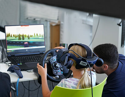 Kind mit Virtual-Reality-Brille vor einem Bildschirm und einem Mann daneben