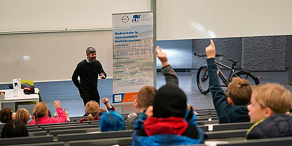 Prof. Christian Rudolph im Hörsaal mit Teilnehmenden der Kinderuni zum Thema Radverkehr, die Kinder melden sich