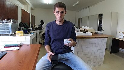 Ein junger Mann hält technische Bauteile in einem Labor mit elektronischer Ausstattung im Hintergrund.