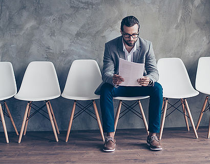 Serious bearded young man in glasses and formal wear is preparing for a meeting, sitting in the hall on chair