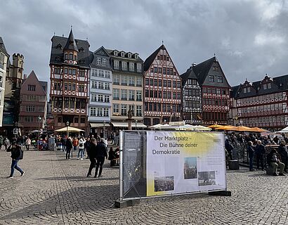 Aufnahme eines Marktplatzes mit bunten Fachwerkhäusern, in der Mitte steht ein Dreiecksmodul der Bauzaunausstellung "Deine Orte der Demokratiegeschichte"