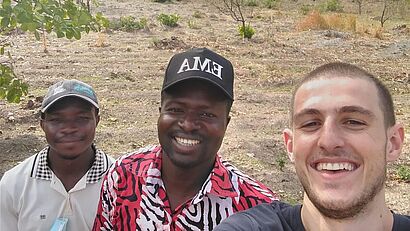 Drei Männer lächeln gemeinsam für ein Selfie in einer ländlichen Außenumgebung.