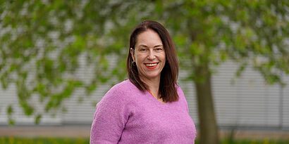 Portraitbild einer Frau mit längerem braunen Haar und pinkfarbenem Pullover vor einem grünblättrigen Baum im Hintergrund