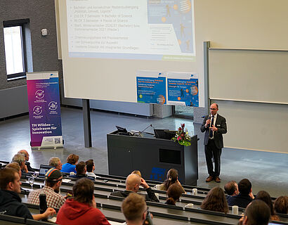 In diesem Bild ist eine Person zu sehen, die etwas vorträgt mit einem Mikrophone in der Hand. Viele Menschen hören in den Reihen zu.