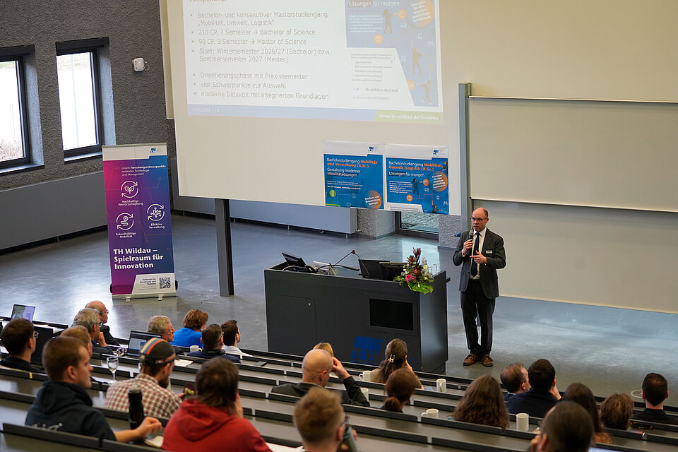 In diesem Bild ist eine Person zu sehen, die etwas vorträgt mit einem Mikrophone in der Hand. Viele Menschen hören in den Reihen zu.