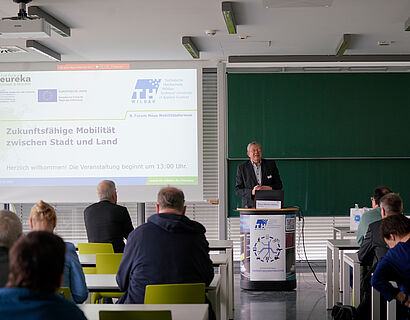 In den Raum wurde von hinten reinfotografiert, es ist eine PowerPoint Präsentation zu sehen und Menschen die an Tischen sitzen.