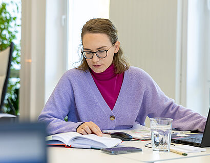 Eine Frau arbeitet konzentriert an ihrem Schreibtisch. Vor ihr sind Bücher aufgeklappt und steht ihr Laptop.