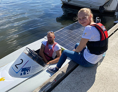 Teilnehmerin der Solarbootregatta sitzt auf dem Steg und hat die Füße im Solarboot auf dem Wasser