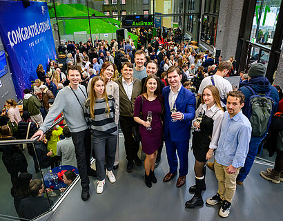 Absolvent*innen stehen für ein Gruppenfoto zusammen, das mit Personen gefüllte Foyer in Halle 17 im Hintergrund