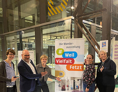 Personen lachen in die kamera und zeigen auf ein Rollup mit der Aufschrift "Weil Vielfalt fetzt"