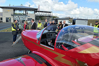 Personen stehen in einer Gruppe zusammen und unterhalten sich, im Vordergrund ist ein Propellerflugzeug zu sehen