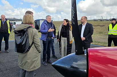 Personen stehen in einer Gruppe auf einem Flugplatz und unterhalten sich, im Vordergrund ist ein Propellerflugzeug zu sehen