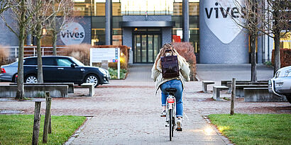 Frau fährt auf einem Fahrrad auf ein Hochschulgebäude mit der Aufschrift Vives zu 