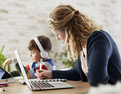 Eine Frau sitzt am Tisch und arbeitet am Laptop. Daneben sitzt ihr Kind mit Kopfhörern und arbeitet ebenfalls an etwas. 