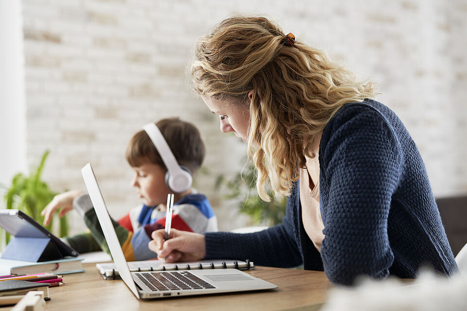 Familie Arbeit Vereinbarkeit Eine Frau sitzt am Tisch und arbeitet am Laptop. Daneben sitzt ihr Kind mit Kopfhörern und arbeitet ebenfalls an etwas.